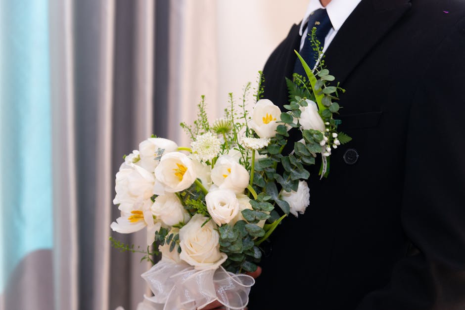 A person dressed in a formal dark suit is holding a bouquet of fresh white flowers, including roses and lisianthus, accented with green foliage such as eucalyptus stems. The bouquet is wrapped with a 