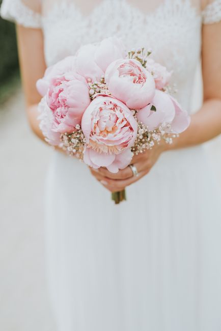 A bride in a white lace dress holding a bouquet of large, pale pink peonies with ruffled petals, accented by small white baby's breath flowers. The bouquet is wrapped with a simple green ribbon, and t
