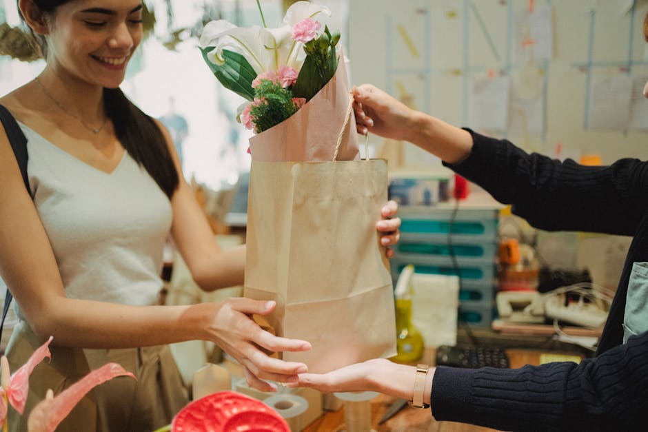 A female florist with dark hair, dressed in a light-colored sleeveless top, is smiling as she hands over a floral bouquet in a brown paper wrapping to a customer. The bouquet contains white lilies, pi