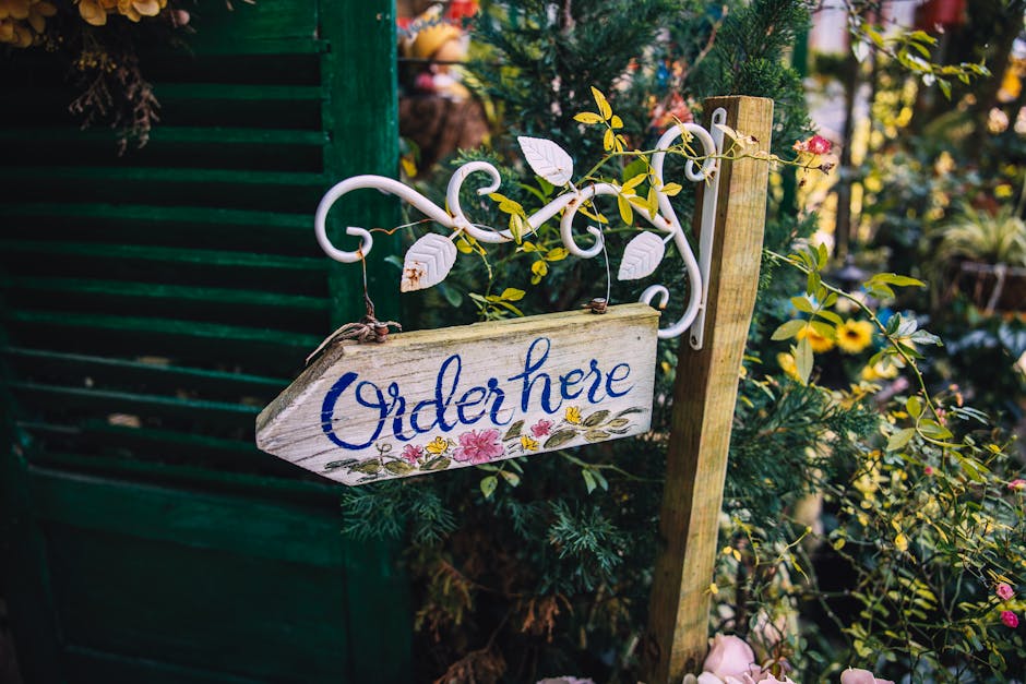 A rustic wooden sign with the word 'Order here' painted in blue cursive, hanging from a white ornate metal bracket attached to a wooden post amidst lush green foliage and vibrant flowers, including ye