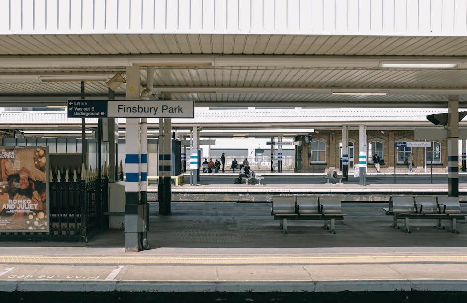 Finsbury Park train station platform exterior with a white overhead canopy, blue and white signage displaying 'Finsbury Park,' several wooden benches, and a few passengers sitting and waiting. In the 