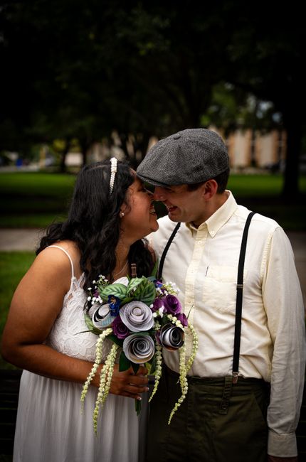 A bride and groom standing closely outdoors in a park with lush green trees and grass in the background, sharing a joyful moment with their foreheads touching. The bride, wearing a white lace wedding 