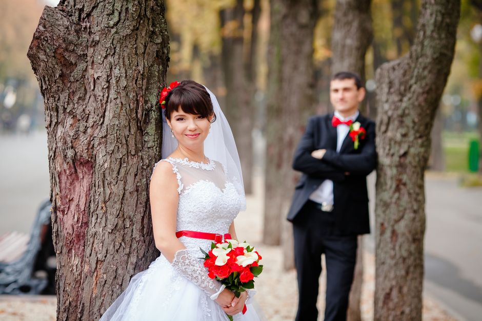 A bride dressed in a white lace wedding gown with a sheer neckline, wearing a red floral headpiece and a white veil, is holding a bouquet of red and white roses with greenery, wrapped in transparent c
